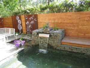 Gartengestaltung mit Sichtschutz Wand aus Holz, Metall-Elementen aus Cortenstahl, einer Natursteinmauer und Wasser in Form eines Naturpools mit Wasserfall.